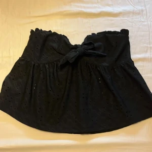 Svart topp med rosett och broderi - Supersöt svart topp med broderade hålmönster och en stor rosett framtill. Toppen har volangkant upptill. Perfekt för dig som gillar detaljer och en feminin stil.