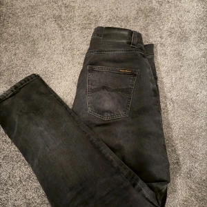 Svarta jeans från Nudie Jeans - Säljer ett par svarta jeans från Nudie Jeans med klassisk femficksdesign och dubbla knappar i midjan. Jeansen har raka ben och diskret logga på bakfickan. Materialet är mjukt denim i bomull och passformen är normal. Perfekta för en clean och stilren look. Modellen är Grim Tim storlek 28/30!