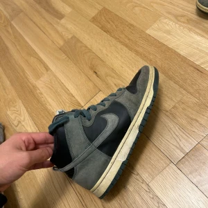 Nike Dunk High i grå och svart mocka - Säljer ett par Nike Dunk High sneakers i en snygg mix av grå mocka och svart läder. Skorna har klassisk Nike Swoosh på sidorna, gröna snören och en vintage-inspirerad sula. Perfekta för dig som gillar streetwear och vill sticka ut med en unik färgkombo.