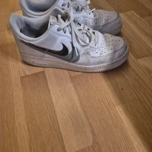 Nike Air Force 1 vita sneakers - Klassiska Nike Air Force 1 sneakers i vitt skinn med svart swoosh-logga på sidan. Lågt skaft, perforerad tå och snörning framtill. Snygg och tidlös modell som passar till det mesta. Perfekt för dig som gillar streetstyle och vill ha en ikonisk sneaker i garderoben.
