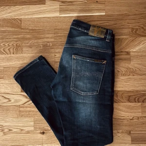 Mörkblå jeans från Nudie Jeans, W31 L30 - Säljer ett par mörkblå jeans från Nudie Jeans med klassisk femficksdesign och snygga slitningar. Jeansen har orangea sömmar, rak passform och är tillverkade i bomull. Perfekta för dig som gillar stilrena och tidlösa jeans.