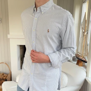 Randig skjorta från Polo Ralph Lauren - Snygg randig skjorta från Polo Ralph Lauren i slim fit. Skjortan är vit med tunna blå ränder, har klassisk krage, knappar framtill och broderad logga på bröstet. Tillverkad i bomull och har långa ärmar.
