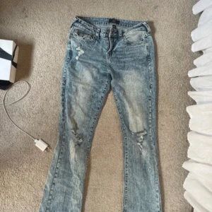 True Religion ljusblå bootcut jeans - Säljer ett par ljusblå bootcut jeans från True Religion med slitningar och snygg tvätt. Jeansen har klassiska bakfickor med lock och röd kontrastsöm samt logopatch bak. Materialet är jeans och passformen är normal med låg midja.