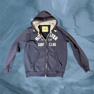 Mörkblå hoodie Hollister L - Snygg mörkblå hoodie från Hollister med broderad vit text på bröstet och mjuk fleecefoder. Tröjan har huva med snörning, dragkedja framtill och två stora fickor. Perfekt för chill dagar och surf vibes.