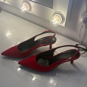 Röda klackar från Pull&Bear - Snygga röda klackar från Pull&Bear med spetsig tå och låg klack. Skorna har en smal rem runt hälen med spänne och är tillverkade i glansigt syntetmaterial. Perfekta för dig som vill sticka ut med en färgklick och elegant siluett.