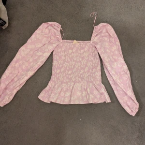 Ljuslila blommig blus med volang - Supersöt ljuslila blus med vita blommor, puffiga långärmar och volang nedtill. Smockad överdel och fyrkantig halsringning.