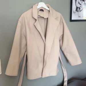 Beige omlott kappa från Missay - Snygg beige kappa från Missay med omlottmodell och bälte i midjan. Jackan har klassisk krage och långa ärmar, perfekt för dig som gillar stilrena och enkla plagg. Materialet är mjukt och ger en chill vibe till din outfit.