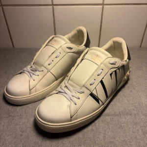 Valentino Garavani skor - Snygga vita Valentino Garavani sneakers i skinn med svart VLTN-logga på sidan och coola nitar runt hälen. Skorna har snörning, rund tå och en silvrig detalj på baksidan. Perfekta för dig som vill sticka ut med exklusiva och trendiga sneakers.