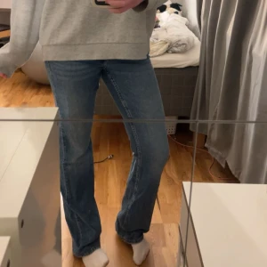 Blå bootcut jeans från Gina Tricot - Säljer ett par klassiska blå jeans från Gina Tricot i storlek 170. Jeansen har låg midja, bootcut-ben och femficksmodell. De är tillverkade i jeansmaterial och har snygga detaljer med knappar på bakfickorna. Perfekta för dig som gillar en tidlös look.