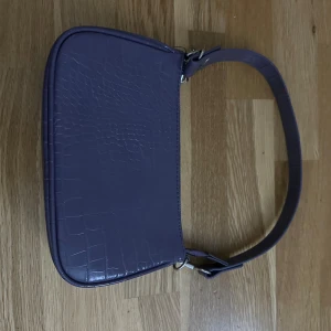 Lila axelväska i krokopräglat skinn - Snygg lila axelväska med croco-mönster och stilren form. Väskan har ett kort axelband och dragkedja upptill. Perfekt för dig som vill ha en trendig accessoar med lite edge. Materialet är skinnimitation med präglad yta som ger en lyxig känsla.
