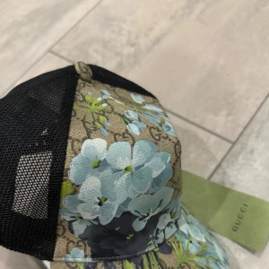 Gucci keps med blå blommor - Snygg keps från Gucci med beige och svart mesh, dekorerad med blå blommor och klassiskt GG-mönster. Framsidan har ett blommigt tryck i ljusblå och gröna toner, medan baksidan är i svart mesh för extra ventilation. Perfekt för dig som vill sticka ut.