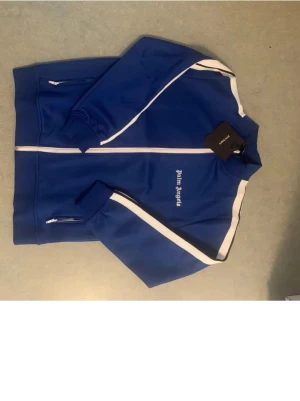 Blå zip hoodie från Palm Angels - Säljer en blå hoodie från Palm Angels med vita ränder längs ärmarna och dragkedja framtill. Tröjan har ribbade muddar, två fickor med dragkedja och logga på bröstet. Perfekt för dig som gillar streetwear och sportig stil.