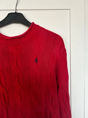 Röd stickad tröja från Ralph Lauren - Säljer en röd kabelstickad tröja från Ralph Lauren med klassisk rund halsringning och broderad marinblå logga på bröstet. Tröjan har långa ärmar och ribbade muddar vid ärmslut och nederkant. Perfekt för kyliga dagar!