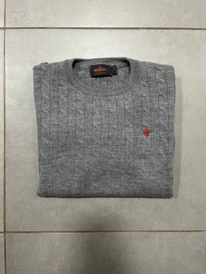 Grå kabelstickad tröja Morris XS - Kabelstickad grå tröja från Morris i 100% merinoull. Klassisk rund halsringning och diskret röd logga broderad på bröstet. Perfekt för dig som gillar stilrena och tidlösa plagg med en modern touch. Står att tröjan är storlek M men passar XS