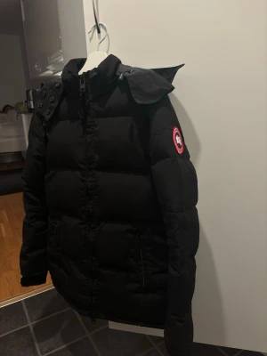  Canada Goose Chelsea Parka - Säljer en svart pufferjacka från Canada Goose med huva och dragkedja framtill. Jackan har det klassiska runda Canada Goose-märket på ärmen och två fickor med dragkedja. Perfekt för kalla dagar och riktigt snygg passform.