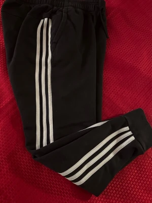 Dam Svarta Adidas mjukisbyxor med vita ränder S  - Svarta mjukisbyxor från Adidas med klassiska vita ränder längs benen och logga på höften. Byxorna har elastisk midja med snörning och är gjorda i mjukt bomullsmaterial. Perfekta för chill eller sport, med normal passform och fickor på sidorna.