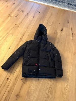 Svart pufferjacka Kings Will Dream - Svart pufferjacka från Kings Will Dream med huva och dragkedja. Jackan har coolt stänkmönster i vitt och rött över hela ytan. Fylld och vadderad för extra värme, perfekt för kalla dagar. Snygg streetstil med normal passform och praktiska fickor.
