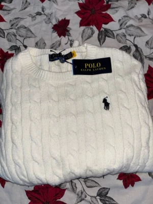 Vit kabelstickad tröja Polo Ralph Lauren - Vit kabelstickad tröja från Polo Ralph Lauren i storlek M. Klassisk design med rund hals och broderad logga på bröstet. Tröjan har långa ärmar och ribbade muddar vid hals, ärmslut och nederkant. Perfekt för dig som gillar stilrena och tidlösa plagg. Priset kan diskuteras. 