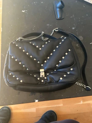 Svart axelväska med nitar och kedja - Säljer en svart axelväska i skinnimitation med quiltad design och silverfärgade nitar i mönster. Väskan har en kedjerem och ett snyggt spänne framtill. Perfekt för dig som vill ha en edgy och trendig look.