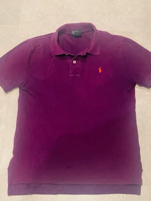 Ralph lauren pike - Säljer en klassisk lila pikétröja från Ralph Lauren med orange broderad logga på bröstet. Tröjan har korta ärmar, krage och knappar framtill. Tillverkad i 100% bomull för en skön och luftig känsla.