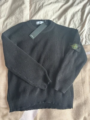 Svart stickad tröja från Stone Island - Säljer en svart stickad tröja från Stone Island med klassisk patch på ärmen. Tröjan har rund halsringning, ribbade muddar och är långärmad. Perfekt för dig som gillar streetwear och vill ha något stilrent och bekvämt.