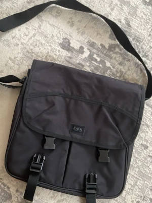 Svart datorväska från ZACK - Säljer en svart datorväska från ZACK med justerbar axelrem och två stora spännen framtill. Väskan har en stilren design och är tillverkad i slitstark polyester, perfekt för att bära laptop och böcker till skolan eller jobbet.