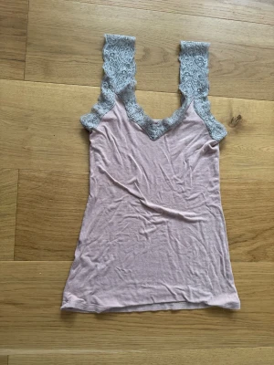 Ljusrosa linne med spetsdetaljer - Säljer ett ljusrosa linne med breda axelband och v-ringning, dekorerat med grå spets längs ringningen och axelbanden. Toppen är ärmlös och har en mjuk, stretchig känsla som gör den bekväm att bära. Perfekt att styla lager på lager eller som den är.