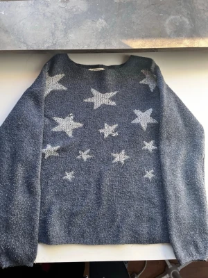 Mörkgrå stickad tröja med stjärnor H&M - Mysig mörkgrå stickad tröja från H&M med vita stjärnor på framsidan. Tröjan har rund halsringning och långa ärmar. Perfekt för dig som gillar en chill och lite lekfull stil.
