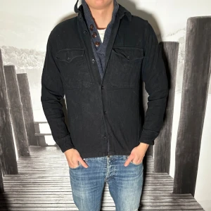 Svart overshirt från Jack & Jones - Säljer en svart overshirt från Jack & Jones med klassisk krage och två bröstfickor med lock. Jackan har knäppning framtill och är tillverkad i ett mjukt bomullstyg. Perfekt att slänga över en t-shirt eller hoodie för en clean look.