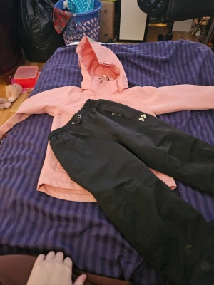 Skalset från från Lager157  - Säljer en ljusrosa skaljacka och med svarta skalbyxor med huva, två dragkedjeförsedda fickor och reflexdetalj på ärmen. Jackan har långa ärmar och stängs med både dragkedja och knapp. Perfekt för blåsiga dagar. Smutsiga pga att man inte ska tvätta så ofta vad jag läst för vattenpelarna.