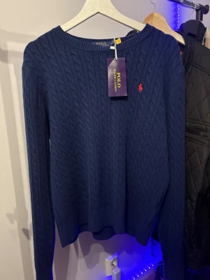 Marinblå kabelstickad tröja Polo Ralph Lauren - Kabelstickad marinblå tröja från Polo Ralph Lauren med klassisk röd logga broderad på bröstet. Tröjan har rund halsringning och långa ärmar, perfekt för lager-på-lager. Tillverkad i mjuk bomull för en skön känsla.