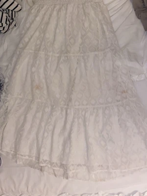 Vit långklänning med spetsdetaljer - Romantisk vit långklänning med vackra spetsdetaljer och volanger. Klänningen har ett diskret geometriskt mönster och resår i midjan för en bekväm passform. Har sminkfläckar på sig 