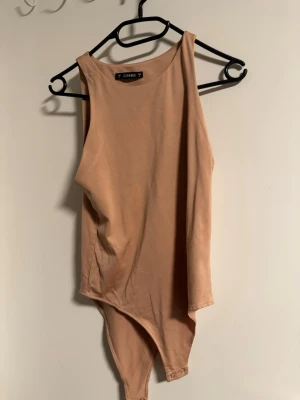 Beige ärmlös bodysuit från Dunnes - Säljer en enkel och stilren beige bodysuit från Dunnes. Modellen är ärmlös med rund halsringning och knäppning i grenen. Perfekt att styla med jeans eller kjol för en clean look.