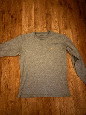 Grå långärmad t-shirt från Carhartt - Säljer en stilren grå långärmad t-shirt från Carhartt i mjuk bomull. Klassisk passform med rund hals och diskret Carhartt-logga broderad i gult på bröstet. Perfekt basplagg för en clean och enkel look.