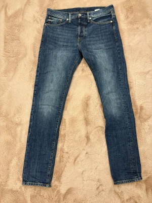 Blå slim straight jeans från H&M - Säljer ett par klassiska blå jeans från H&M i modellen slim straight. Jeansen har fem fickor, normal midja och snygga slitningar för en avslappnad look. Tillverkade i tåligt denim med kontrastsömmar och knappgylf.