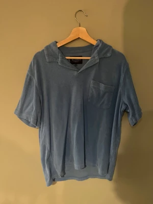 Blå frottétopp från Bondelid  - Mjuk blå frottétopp från Bondelid med korta ärmar och en avslappnad krage. Toppen har en bröstficka och en loose passform som ger en chill vibe. Perfekt för sommarens lata dagar eller när du vill ha något bekvämt och svalt.