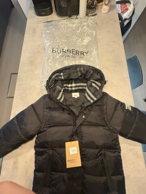 Svart pufferjacka och väst från Burberry L - Säljer en svart pufferjacka från Burberry i storlek L. Jackan har huva med grått och vitt rutigt foder, stora fickor med dragkedja och Burberry-logga på ärmen. Perfekt för kalla dagar och riktigt snygg streetstil.