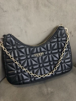 Svart axelväska från Guess - Säljer en svart axelväska från Guess med guldfärgad kedja och dragkedja. Väskan har ett elegant geometriskt mönster, Guess-logga framtill och innerfack med dragkedja. Perfekt storlek för det viktigaste. Använd endast ett fåtal gånger, precis som ny!
