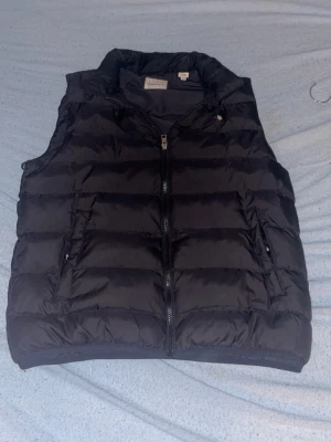 Svart dunväst från GANT - Säljer en svart dunväst från GANT med hög krage och dragkedja framtill. Västen har två sidofickor med dragkedja och quiltad design. Perfekt för lager på lager under höst och vinter. Klassisk och stilren look. Den har ett litet hål men fungerar bra kan sänka ner priset vid snabb affär ny pris 1000