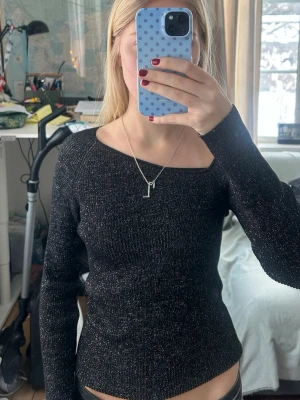 Glittrig svart topp  - Säljer en svart stickad topp från NORR med glittriga inslag. Toppen har lång ärm och en snygg asymmetrisk halsringning som ger en unik look. Materialet är stretchigt och sitter tajt mot kroppen. Perfekt för dig som vill sticka ut lite extra på festen.