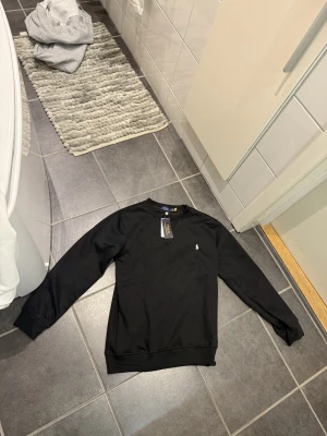 Svart tröja från Polo Ralph Lauren - Säljer en svart långärmad tröja från Polo Ralph Lauren med klassisk rund hals och den ikoniska vita logon broderad på bröstet. Tröjan är i mjuk bomull och har ribbade muddar vid ärmslut och nederkant. Perfekt för en clean och stilren look. Aldrig använts!