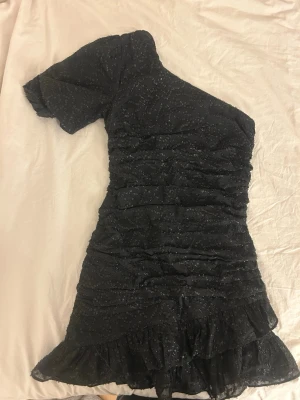 Svart glittrig one shoulder klänning - Supersnygg svart kortklänning från MANGO med glittriga detaljer och volangkant nertill. One shoulder-design med en ärm och tight passform. Perfekt för dig som vill sticka ut med en unik look. Materialet känns lätt och har en skimrande finish.