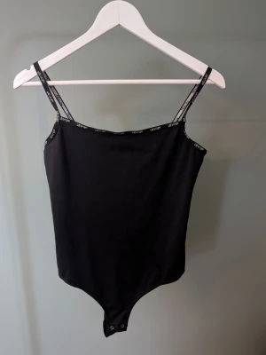 Svart bodysuit från Levi's - Svart bodysuit från Levi's med tunna axelband och logotypdetaljer längs kanterna. Modellen är ärmlös och har tryckknappar i grenen. Tillverkad i mjuk bomullsmix som sitter tight och bekvämt.