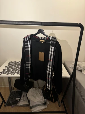 Svart hoodie med rutig huva Burberry - Svart hoodie från Burberry med dragkedja och snörning. Insidan av huvan och öppningen har klassiskt rutigt mönster i svart, vitt och rött. Tröjan har långa ärmar, fickor framtill och en broderad logga på bröstet. Perfekt för dig som gillar stilrena streetwear-plagg.  Finns i storlek S,M och L