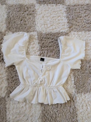 Vit blus med snörning och volang - Supersöt vit blus från Gina Tricot med puffiga korta ärmar, snörning framtill och volangkant nedtill. Tillverkad i ett lätt, strukturerat tyg som ger en luftig känsla. Perfekt för sommarens alla tillfällen!