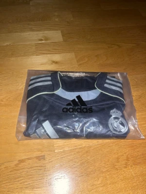 Adidas Real Madrid matchtröja svart - Säljer en officiell Real Madrid matchtröja från Adidas i mörkblått med silverdetaljer och klubbmärke på bröstet. Tröjan har korta ärmar, rund hals och klassiska Adidas-ränder på axlarna. Materialet är lätt och andas, perfekt för fotboll eller träning.
