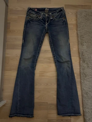 True Religion bootcut jeans  - Säljer ett par lågmidjade blå jeans från True Religion med bootcut passform och kontrasterande vita sömmar. Jeansen har detaljerade fickor med lock och stora silverknappar, samt en tydlig logopatch bak. Säljer dessa jeans för att jag har slutat använda de. De är lite slitna där jeansen slutar men annars fel fria. Köpta för 1200kr.