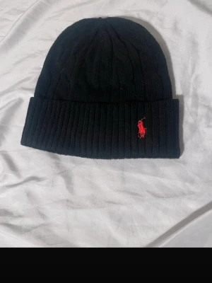 Svart mössa från Polo Ralph Lauren - Svart ribbstickad mössa från Polo Ralph Lauren med klassisk röd broderad logga framtill. Mössan är i mjuk ull och har en vikt kant för extra stilpoäng. Perfekt accessoar för kyliga dagar.