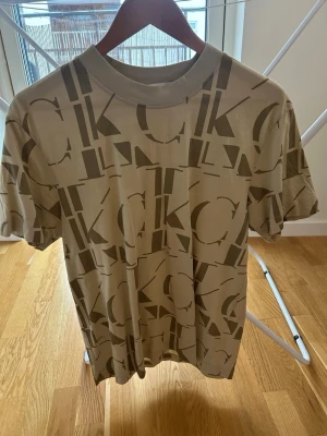Beige CK t-shirt från Calvin Klein Jeans - Säljer en beige t-shirt från Calvin Klein Jeans med stort CK-logomönster över hela plagget. T-shirten har rund hals, korta ärmar och är gjord i mjuk bomull. Perfekt för dig som gillar streetwear och vill sticka ut lite extra.
