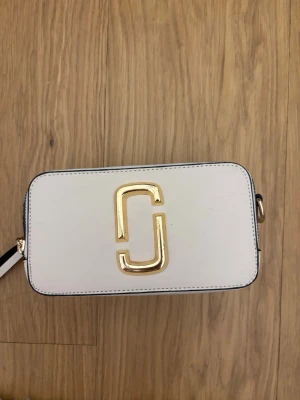 Vit axelväska från Marc Jacobs - Säljer en stilren vit axelväska från Marc Jacobs med rektangulär form och stort guldfärgat logotyp-spänne på framsidan. Väskan har svarta detaljer och ingen axelräm. Perfekt för dig som vill ha en snygg och enkel accessoar.